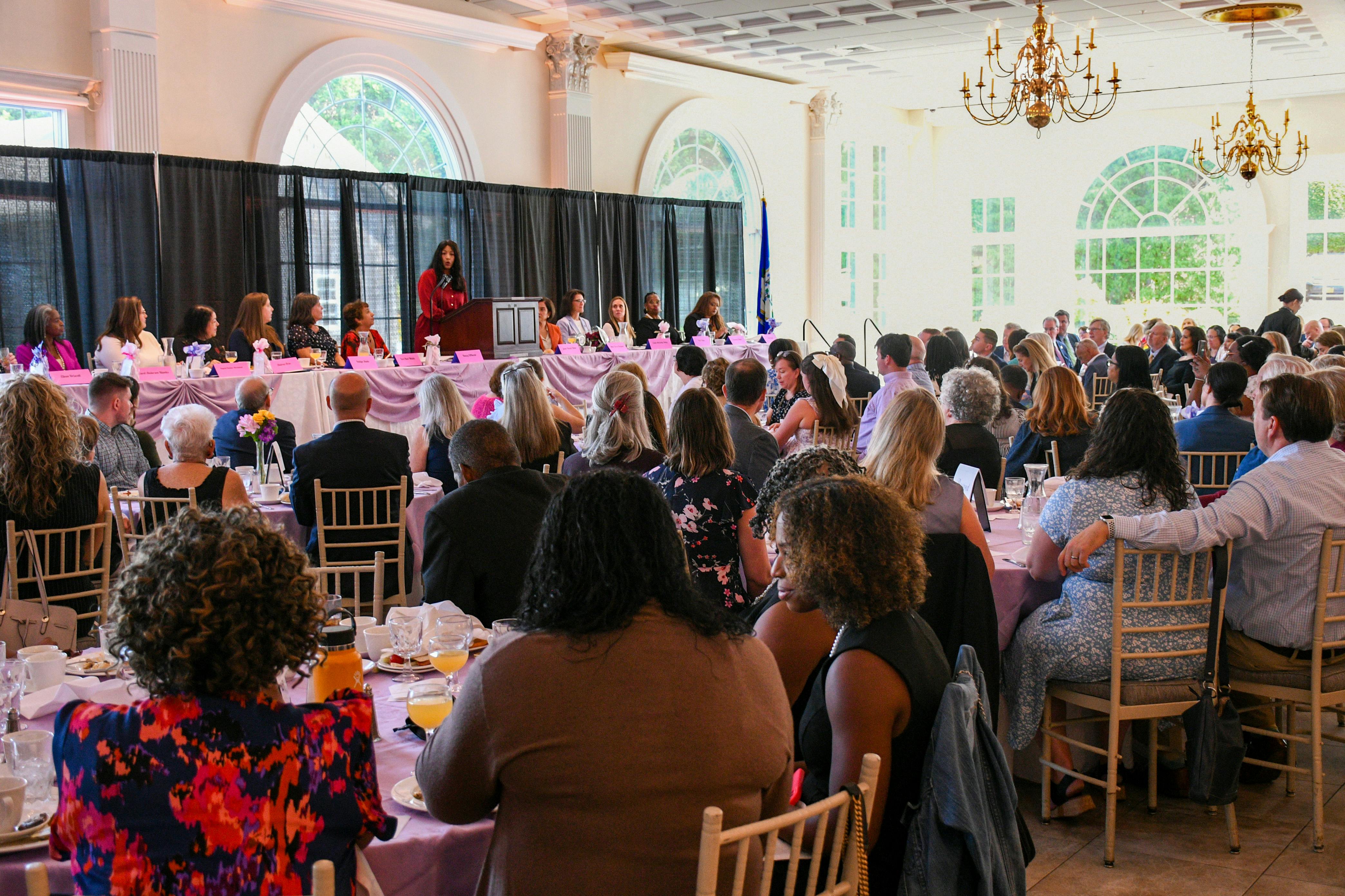 Guests listen to keynote address at WLB. Panelists and speakers are at a dias with a black curtain behind them