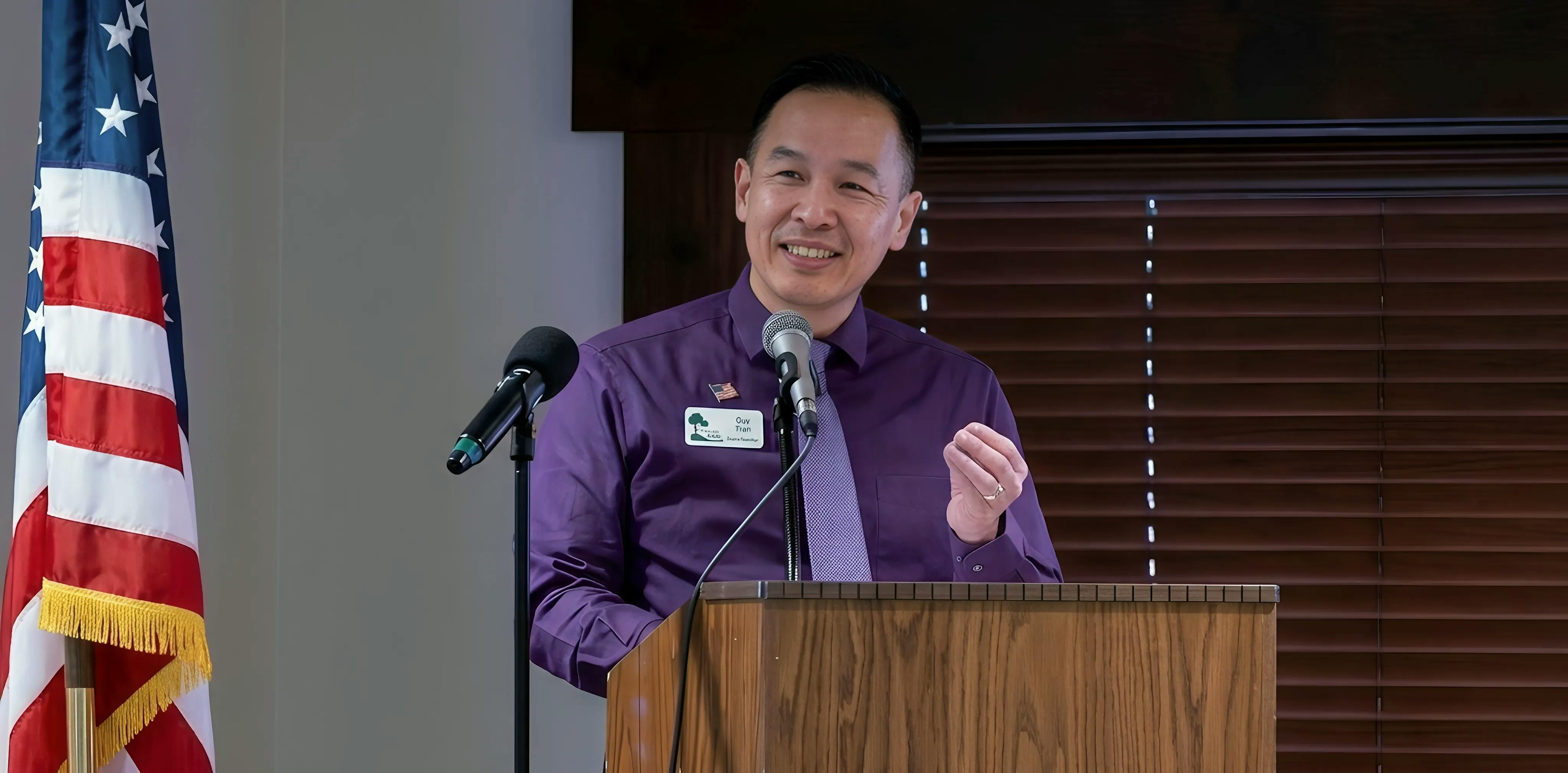 Candidate Dr. Quy Tran wearing purple speaking into a microphone at a lectern. He is smiling and wearing his Rec and Park name badge.. 