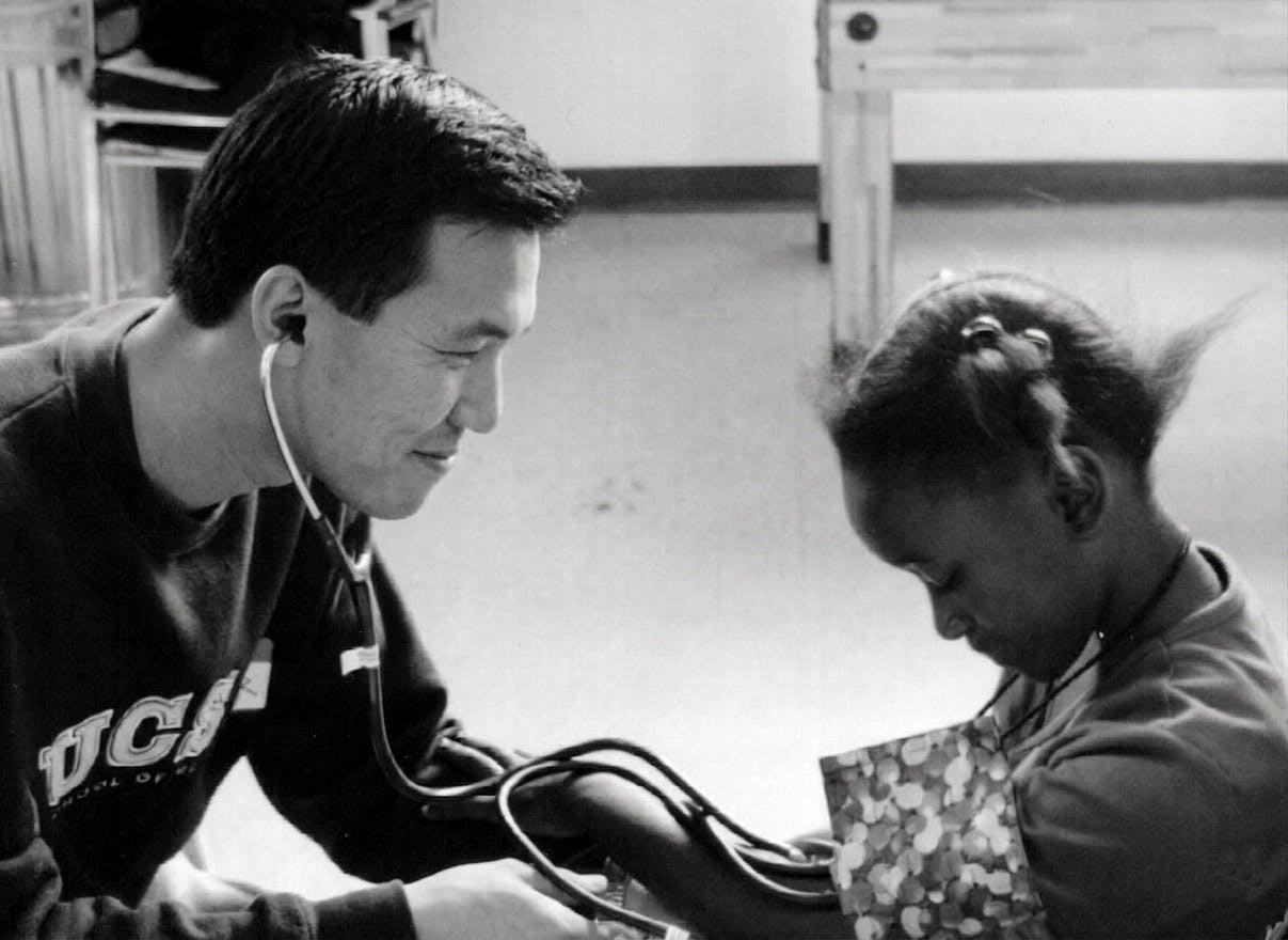 Candidate Dr. Quy Tran on left with stethoscope helping a child patient on right. Image is in black and white.