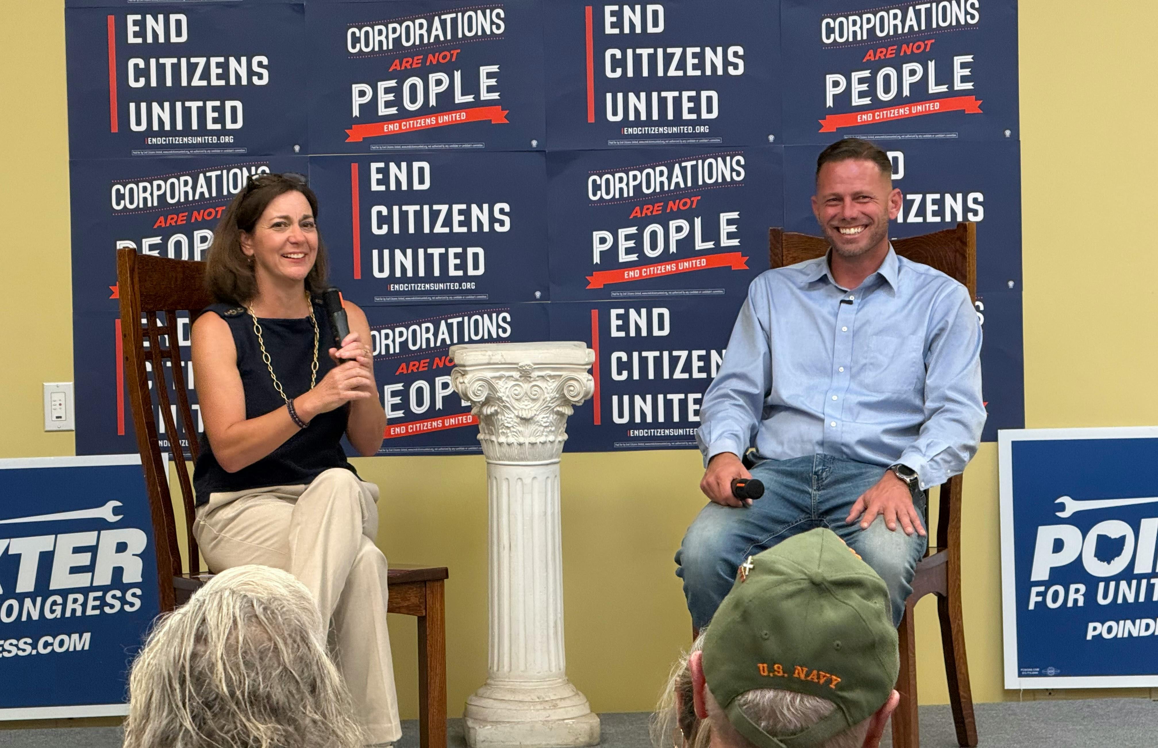 ECU President Tiffany Muller and congressional candidate Brian Poindexter are seated next to each other smiling. Behind them are placards with End Citizens United and Corporations are not people. On both sides, there are signs for Poindexter for Congress. The back of heads of members of the crowd are in the foreground.