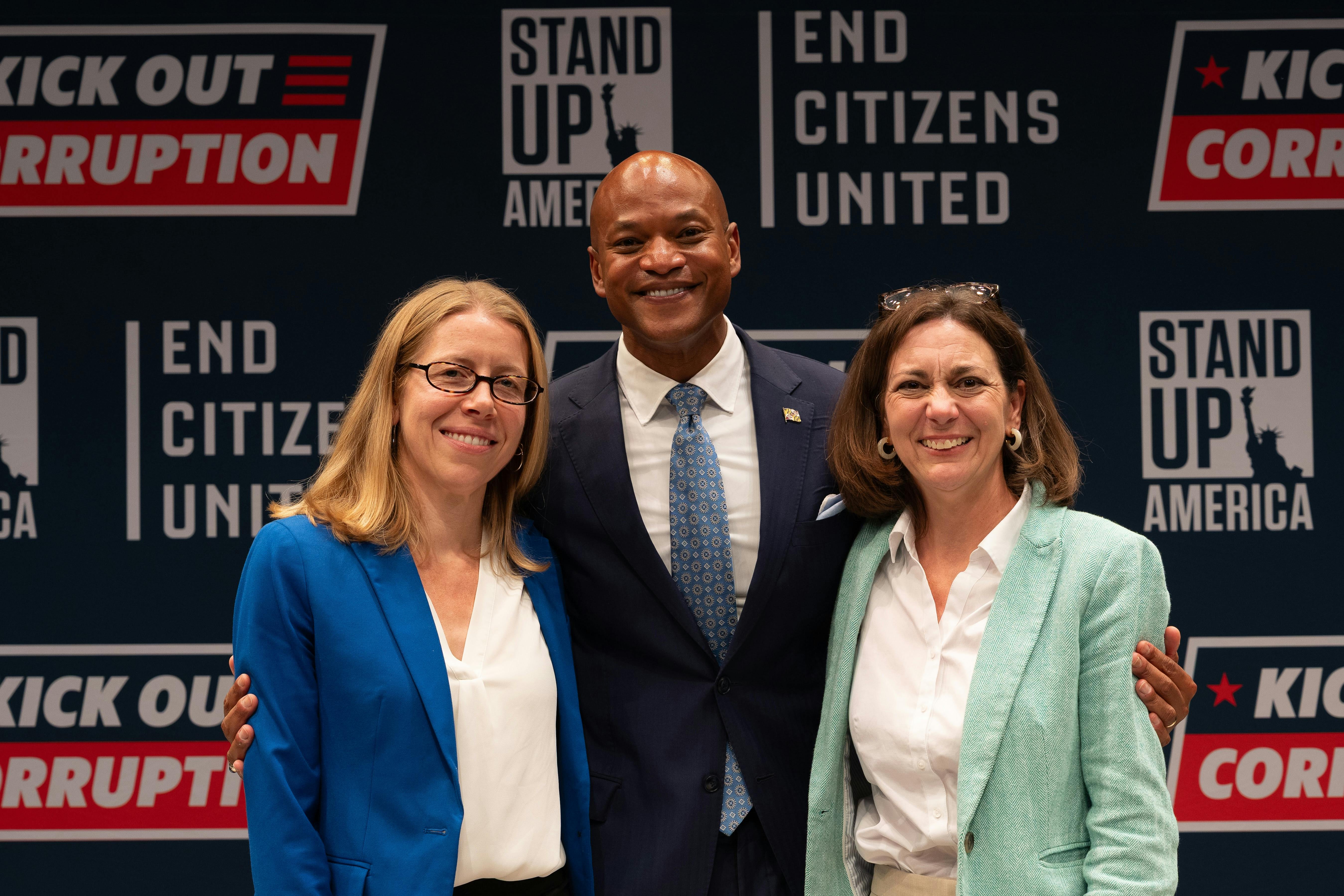 Left to Right: Stand Up America Executive Director Christina Harvey, Governor Wes Moore, and End Citizens United President Tiffany Muller smile for a photo. Behind them is a wall covered with signs for Stand Up America, End Citizens United, and the Kick Out Corruption tour. 