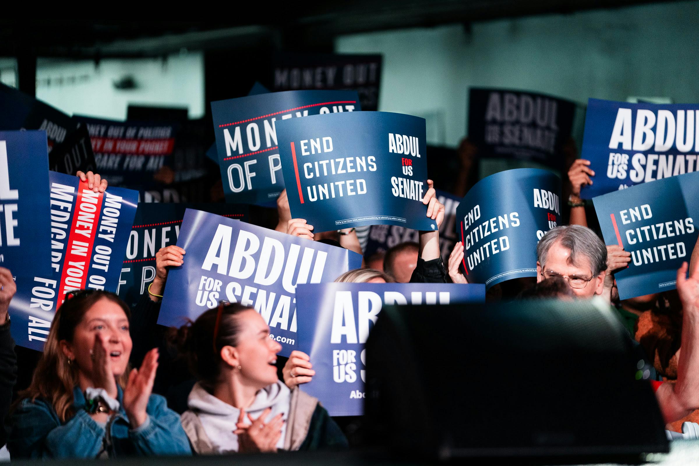 Crowd of people applauding carrying End Citizens United and Abdul for Senate signs.
