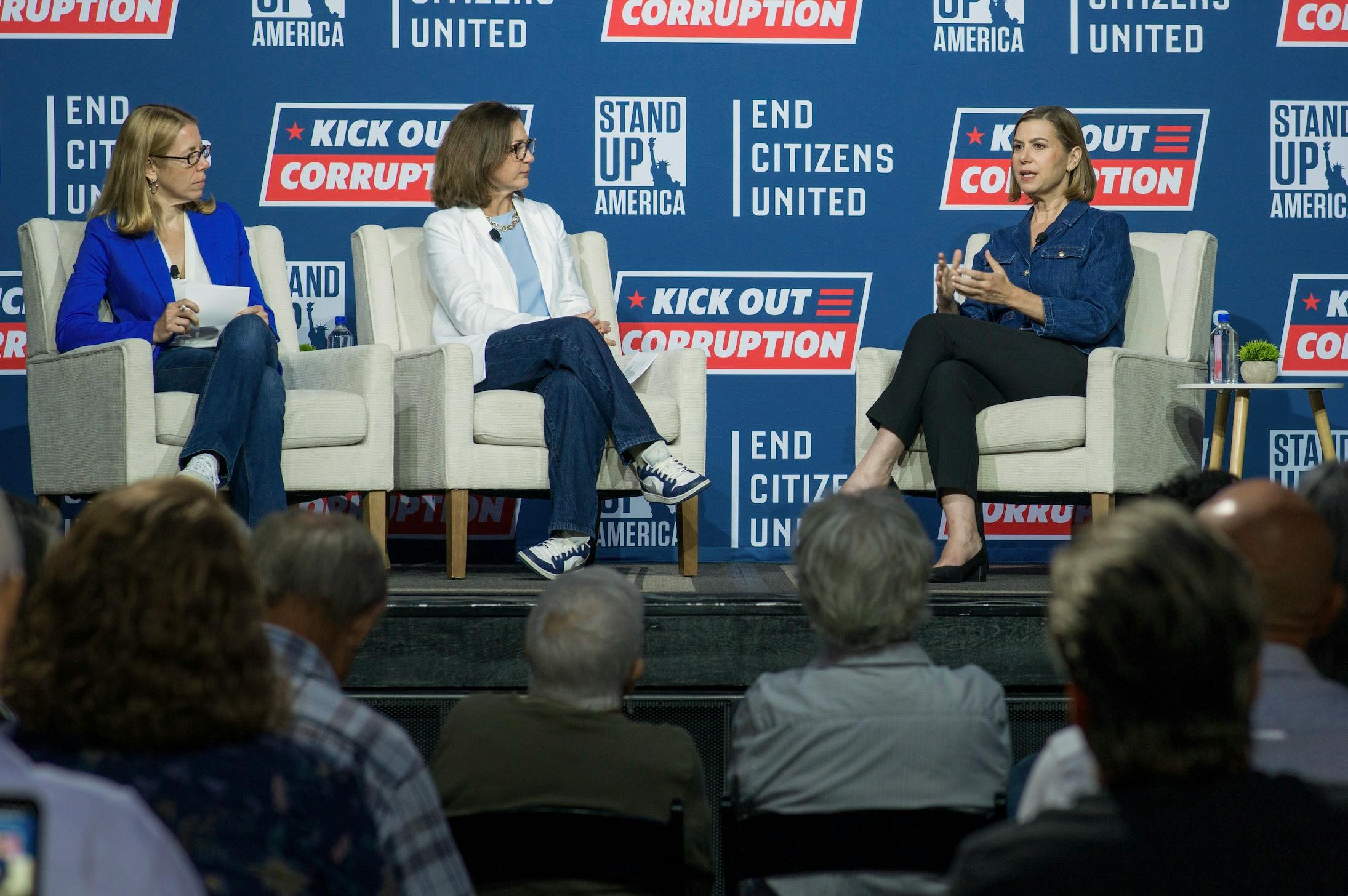 Stand Up America Christina Harvey, ECU President Tiffany Muller, and Senator Elissa Slotkin sit on stools on a platform stage talking. Senator Slotkin holds a mic she is speaking into. Signs behind them read Stand Up America, End Citizens United, Kick Out Corruption.