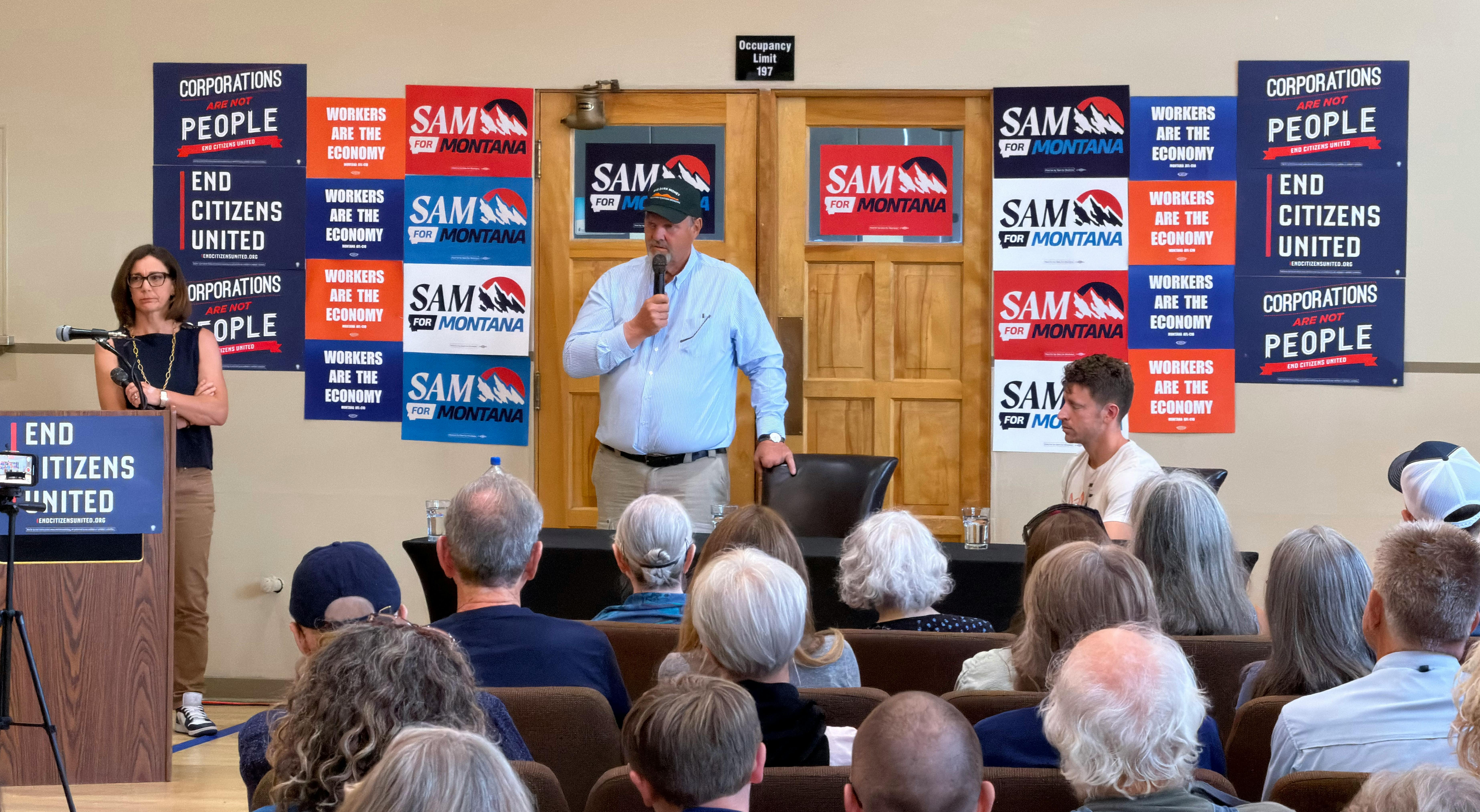 Tiffany Muller, Jon Tester, and Sam Forstag in front of crowd at town hall. Jon Tester is speaking, audience is in the foreground. 