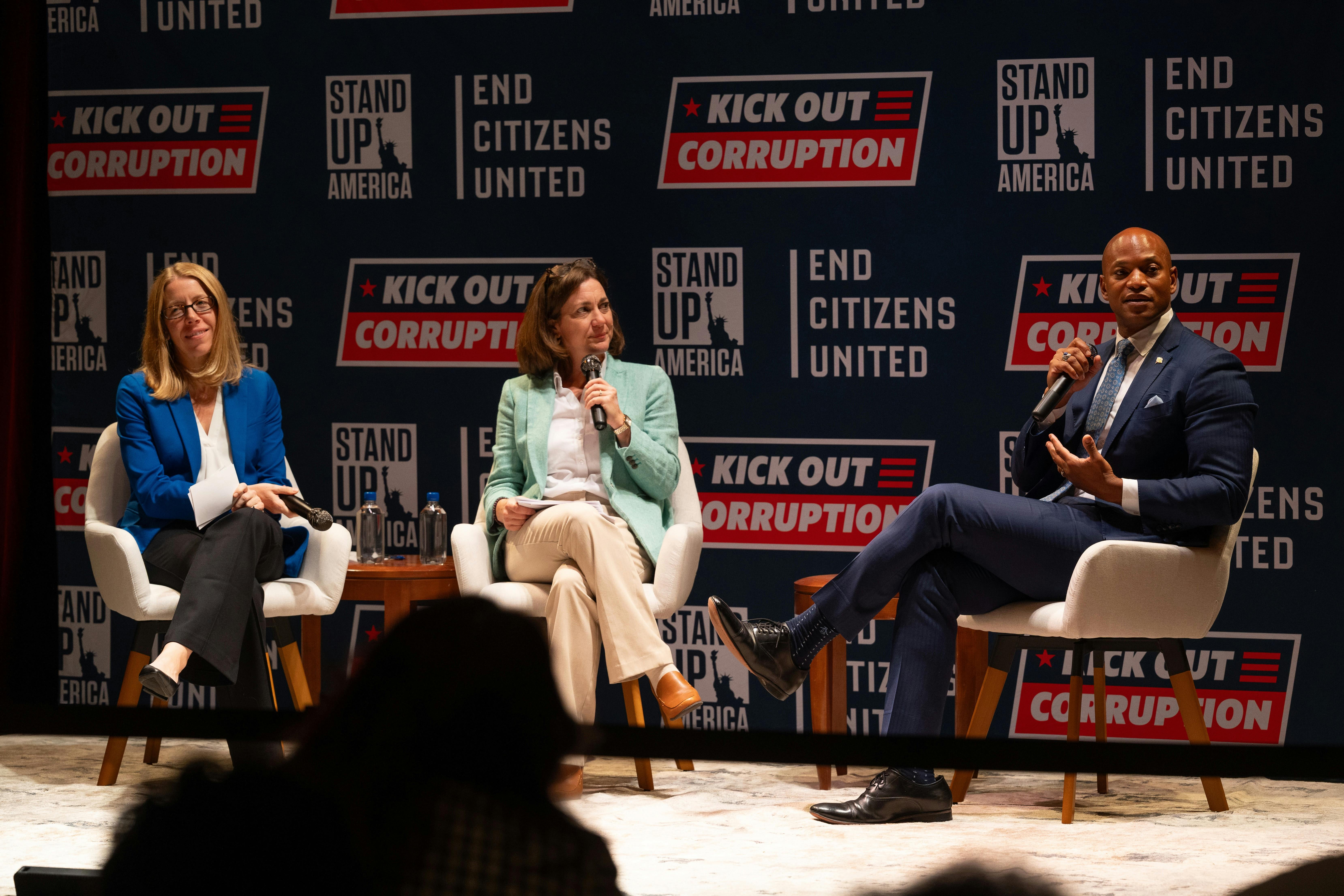 Stand Up America Executive Director Christina Harvey, End Citizens United President Tiffany Muller, and Governor Wes Moore sit on chairs on a stage. Governor Moore is talking to the audience. Behind them is a wall with logos for Stand Up America, End Citizens United, and the Kick Out Corruption tour.