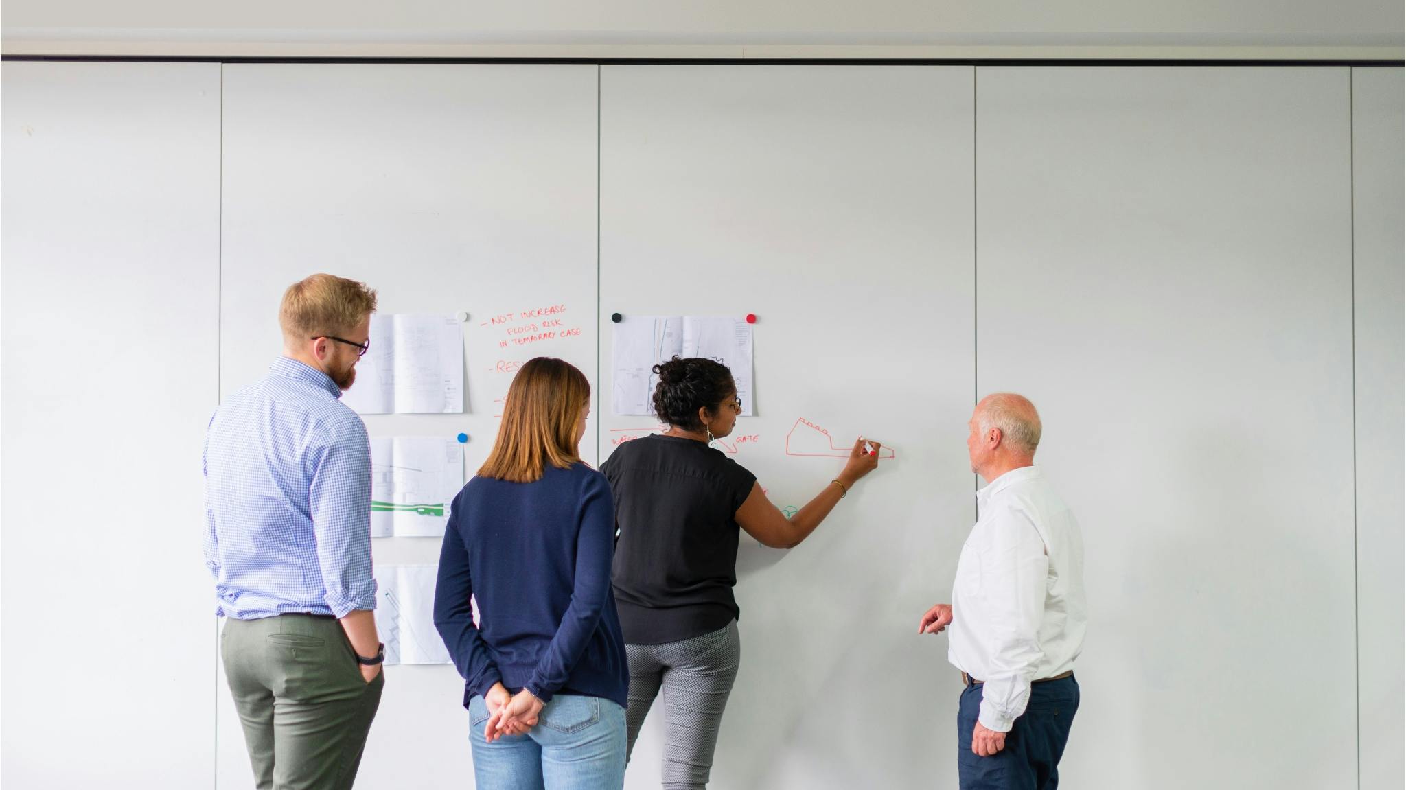 A diverse group of four gathers at a whiteboard covered in notes and diagrams, working through a plan together.