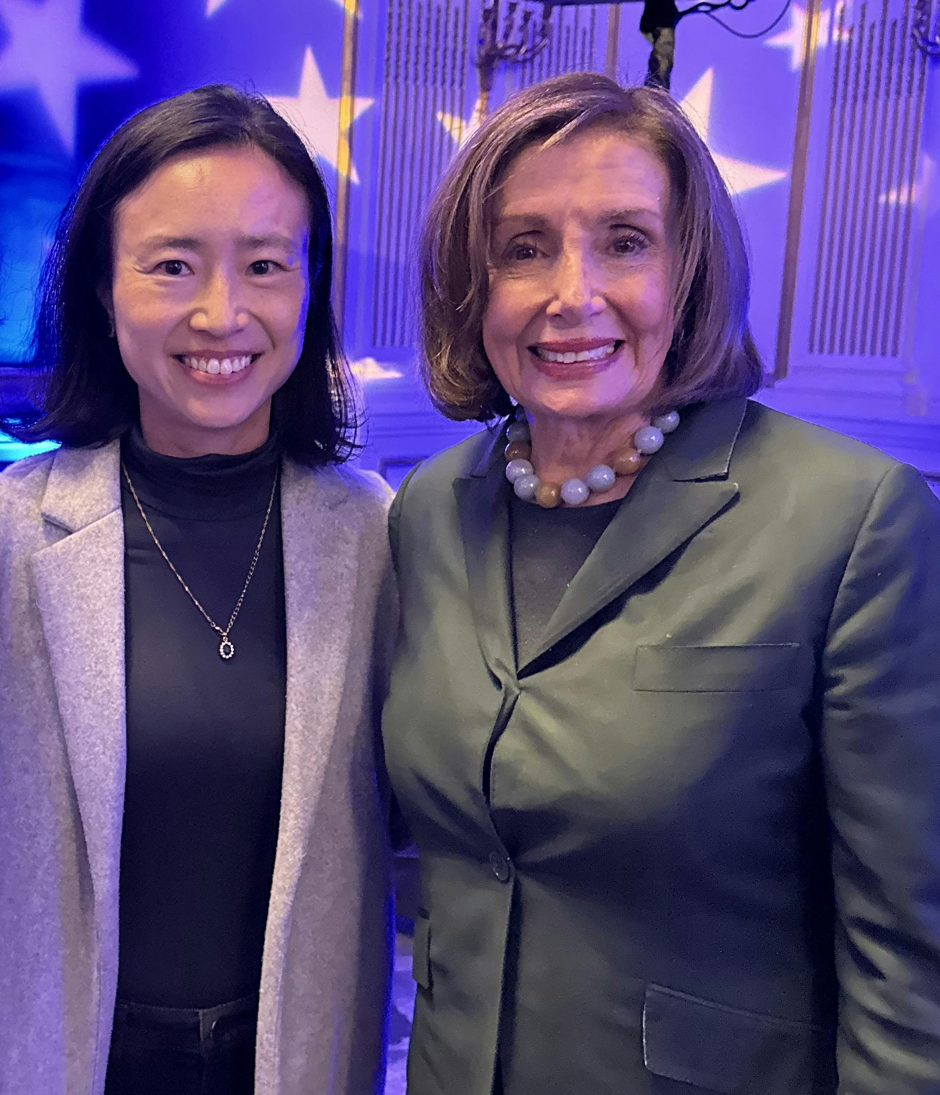 Connie Chan smiling while standing beside Speaker Nancy Pelosi at an indoor event with star-shaped lights projected on the wall.