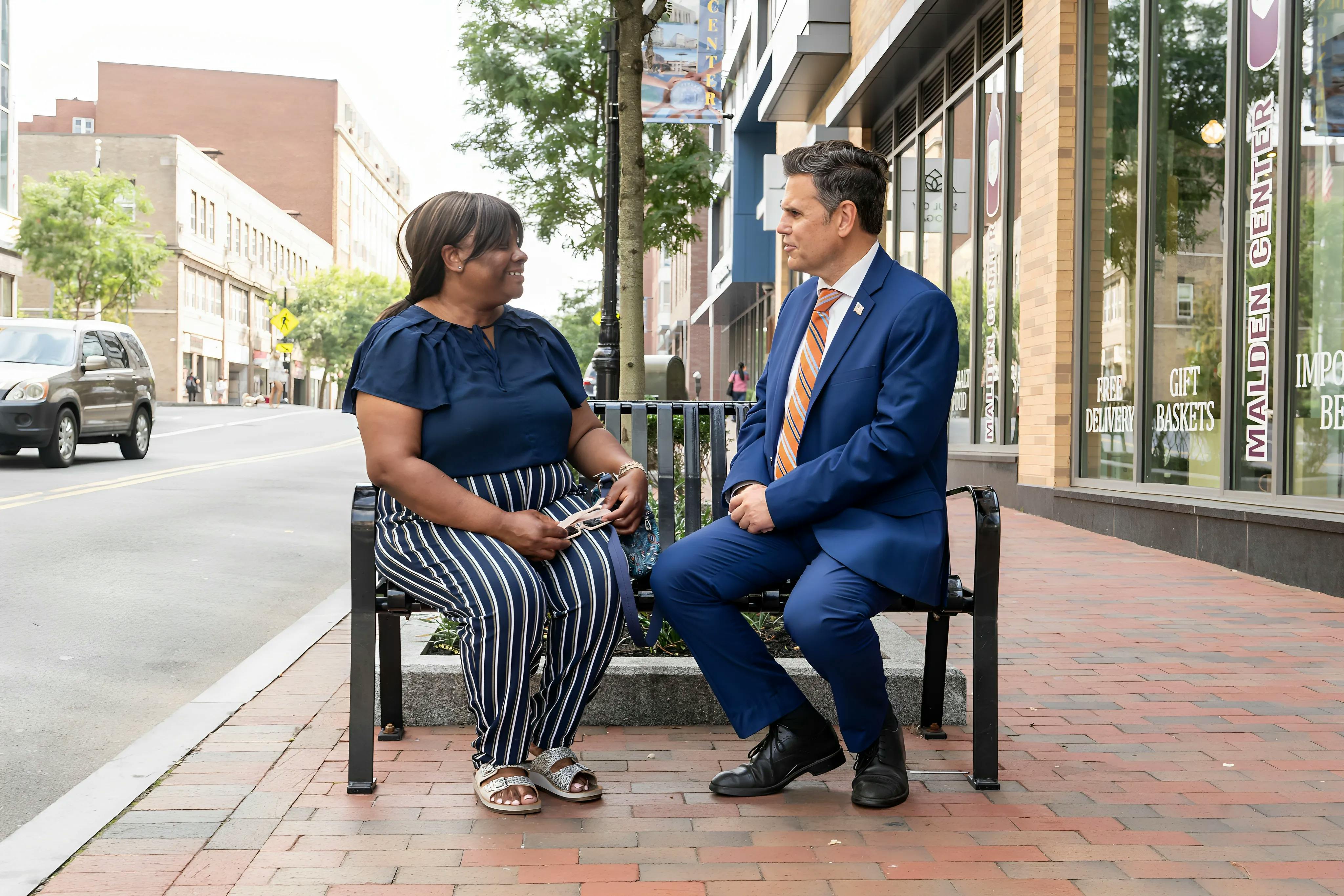 Carline Louis, a Black woman in striped pants and a blouse sitting with Mayor Christenson on a bench in Malden Center.