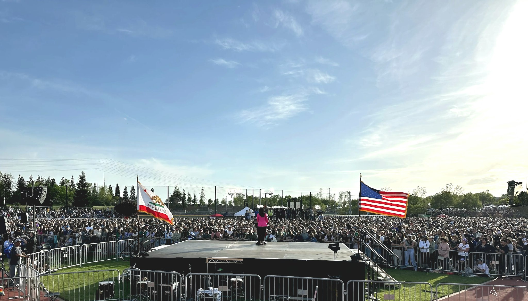 Jane Kim stands on stage in a pink blazer, speaker to a crowd of tens of thousands of people. To her left is the California state flag, and to her right the American flag.