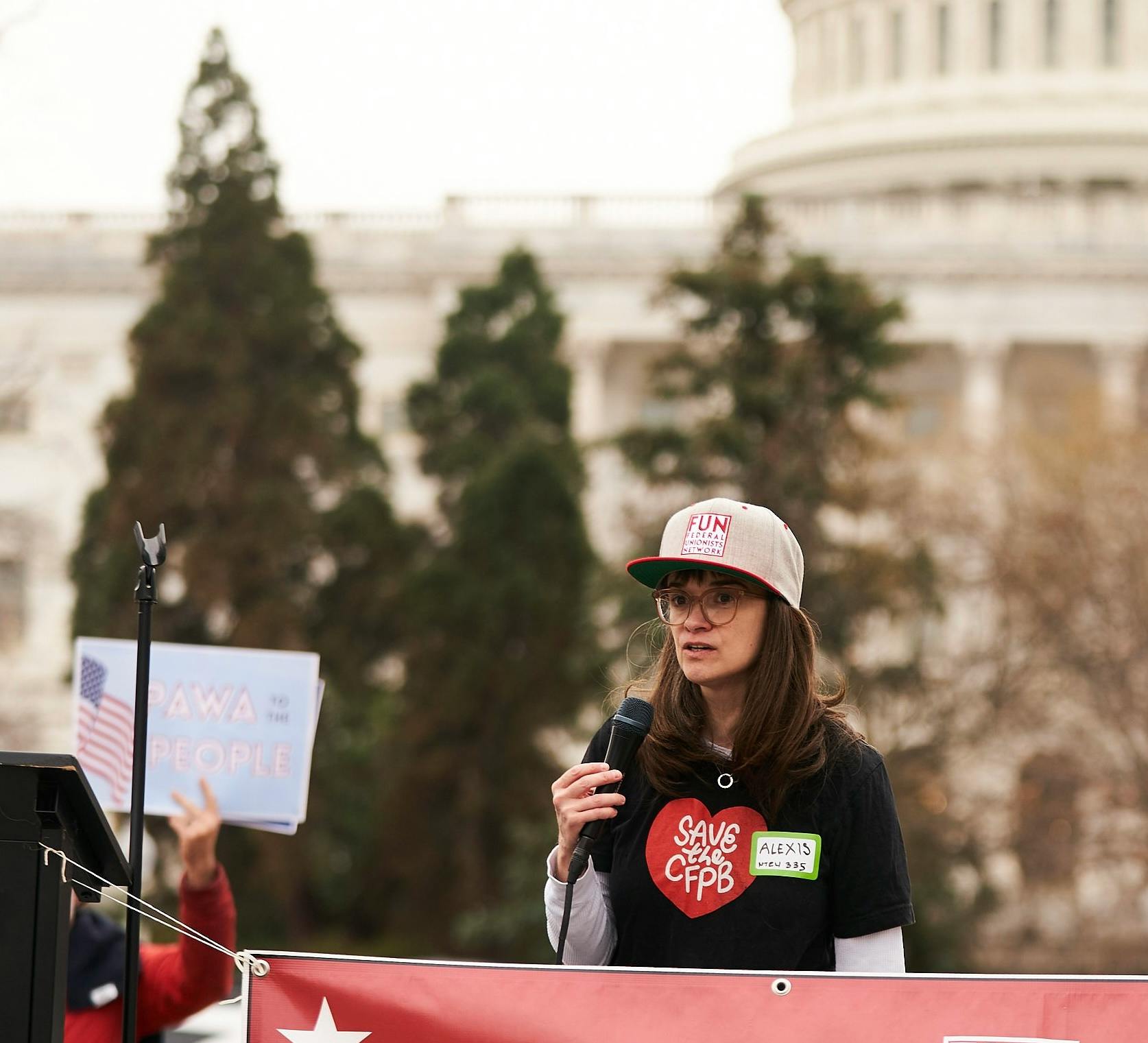 Alexis Goldstein con una gorra de béisbol que dice "FUN: Federal Unionists Network (Red de Sindicalistas Federales)" y una camiseta negra con un corazón rojo que dice "Save the CFPB (Proteja la Oficina de Protección Financiera del Consumidor".
