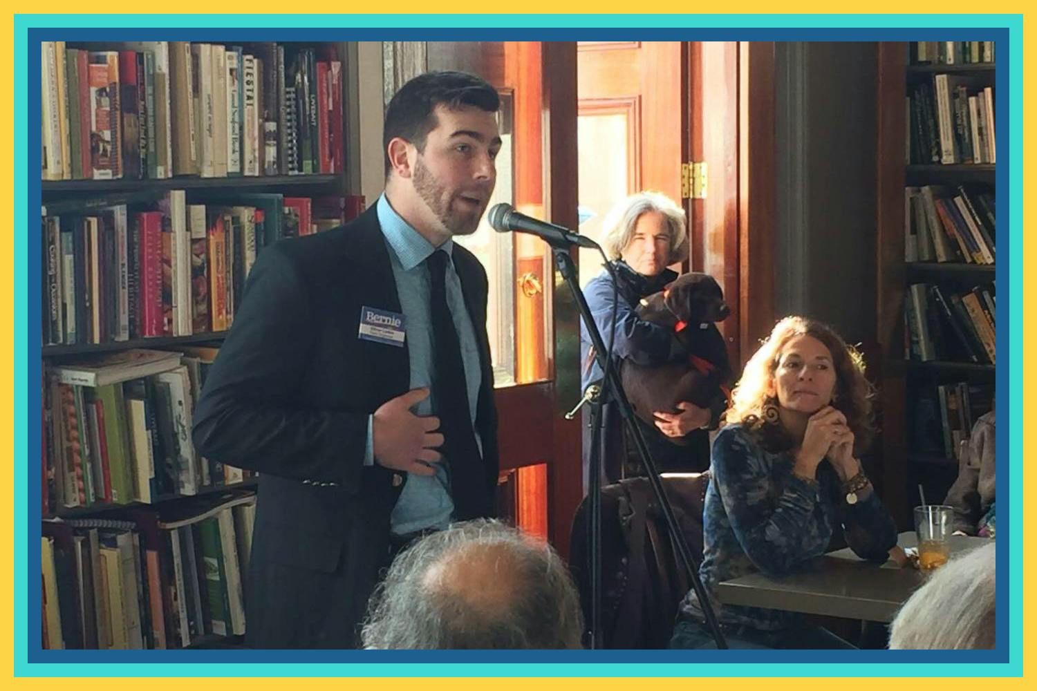 A man, Oliver Larkin, in a suit and tie speaks into a microphone at a bookstore at a campaign event.