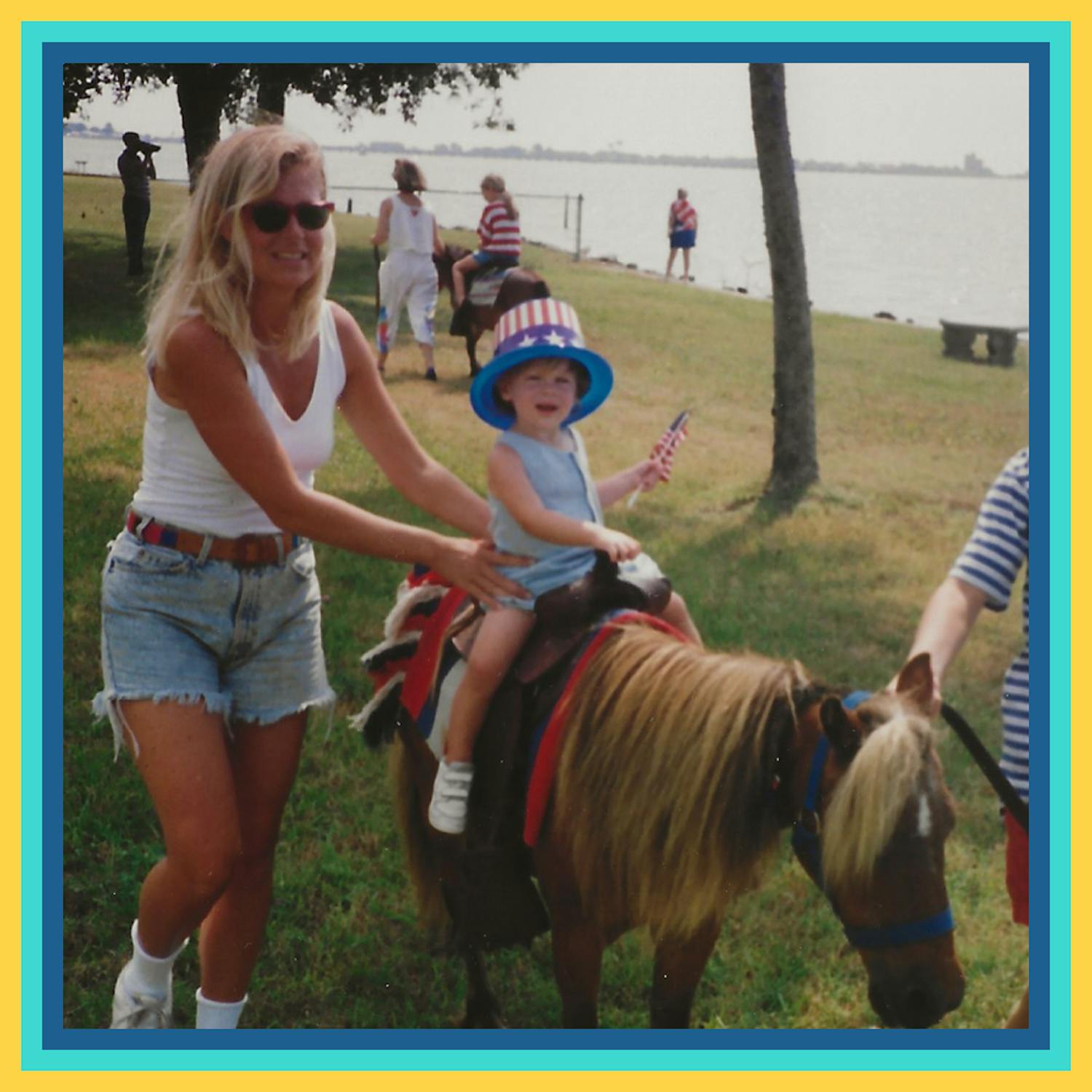 Oliver, as a child, riding a small, brown miniature horse, holding an American flag with an American flag top hat, with his mom, Margot standing by his side, holding onto him in a sunny outdoor setting.