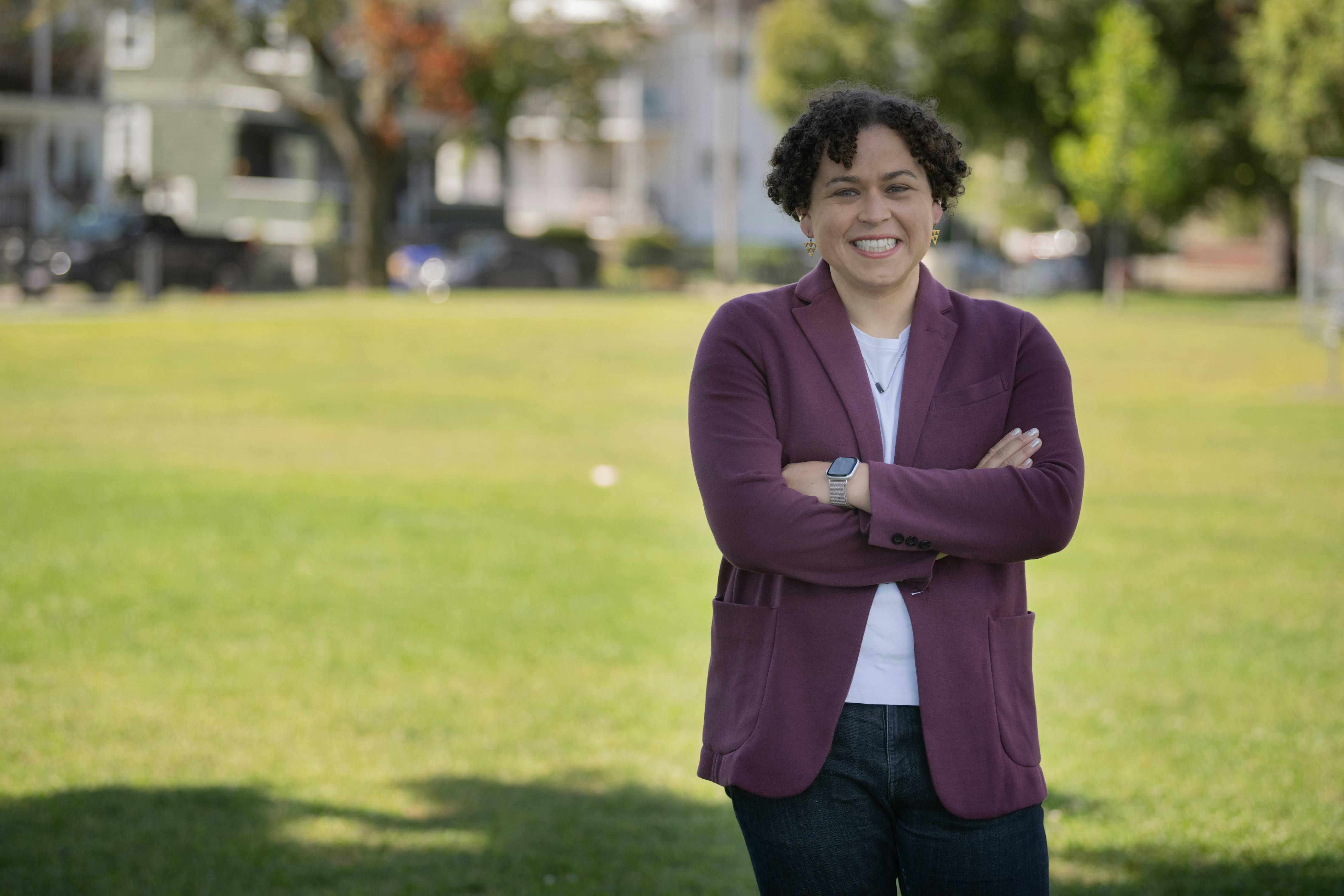 Carey stands in a park, arms crossed and smiling, facing the camera