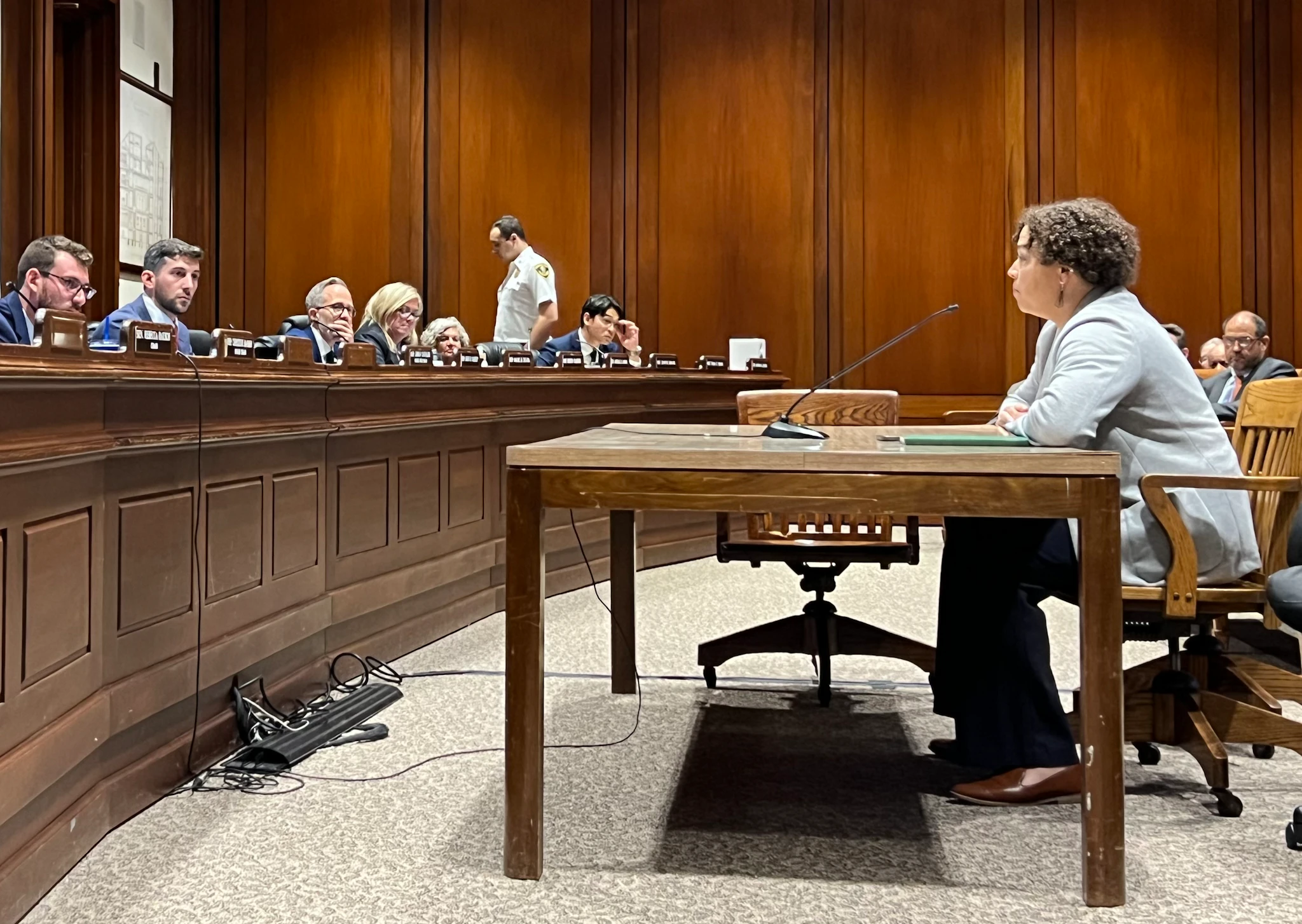 Side shot of Carey sitting at a table, testifying before a legislative panel in a wood-paneled statehouse commitee room