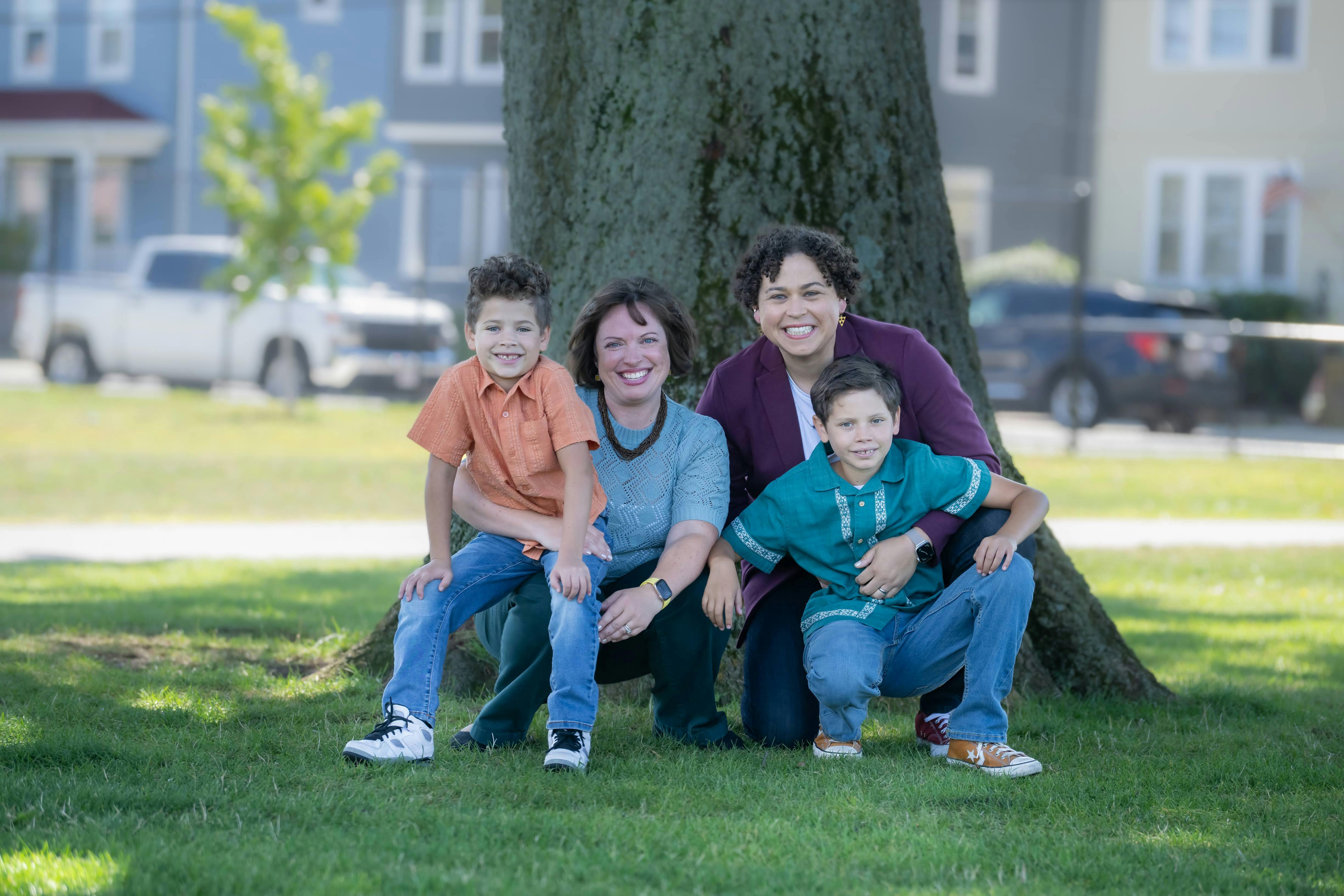 Carey's family - spouse and two young kids - crouch down while smiling for a photo in the park