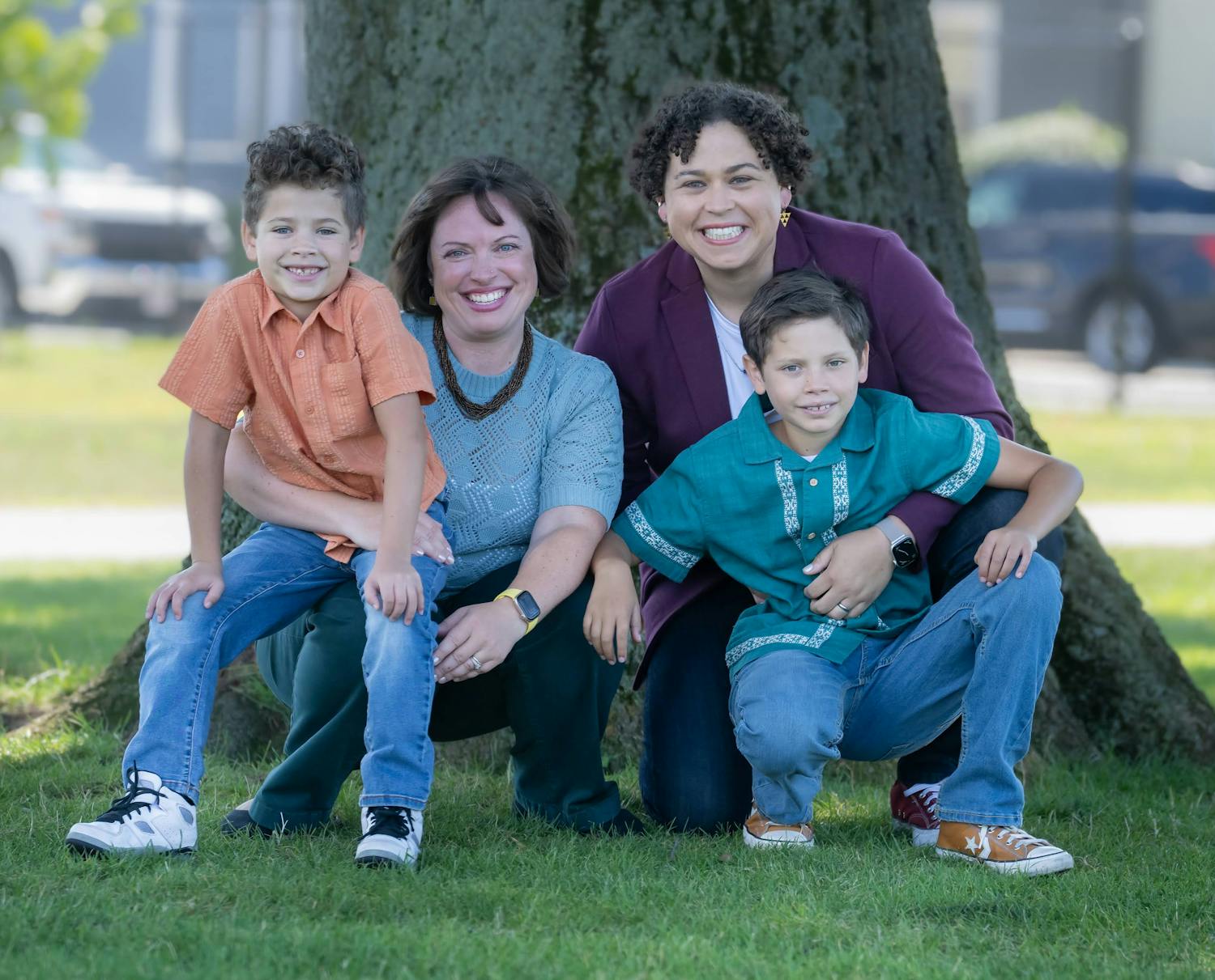 Carey's family - spouse and two young kids - crouch down while smiling for a photo in the park
