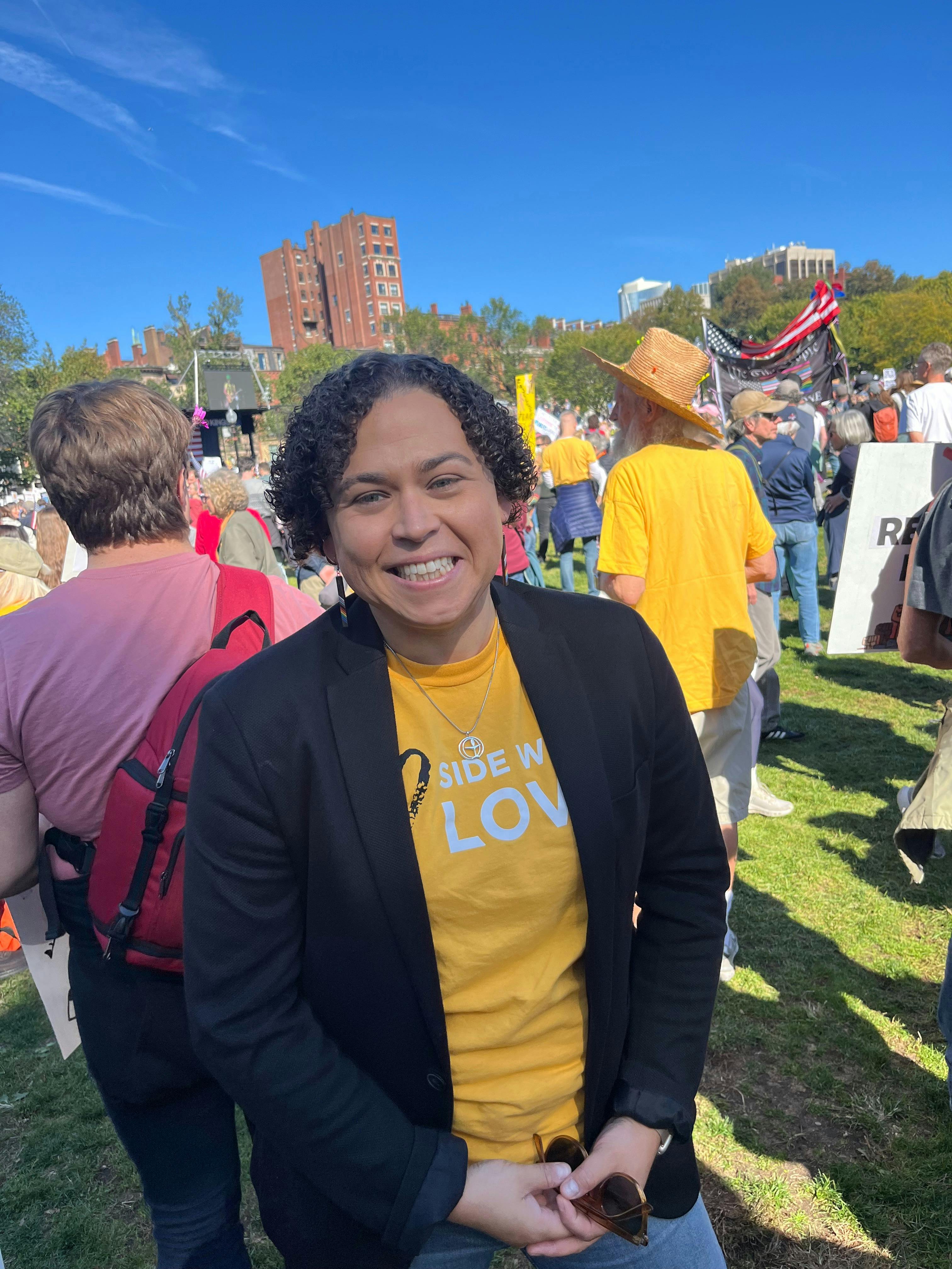 Carey is smiling standing in a crowd, wearing a yellow Side With Love shirt under a black blazer