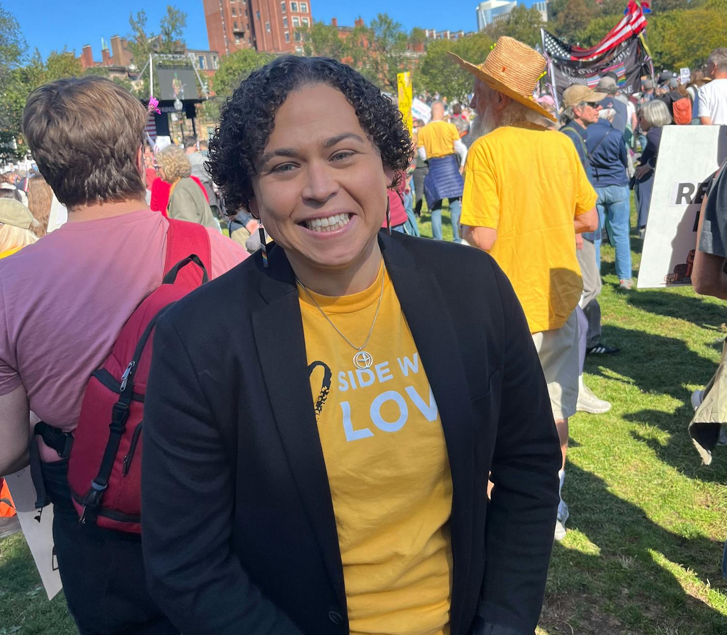 Carey is smiling standing in a crowd, wearing a yellow Side With Love shirt under a black blazer