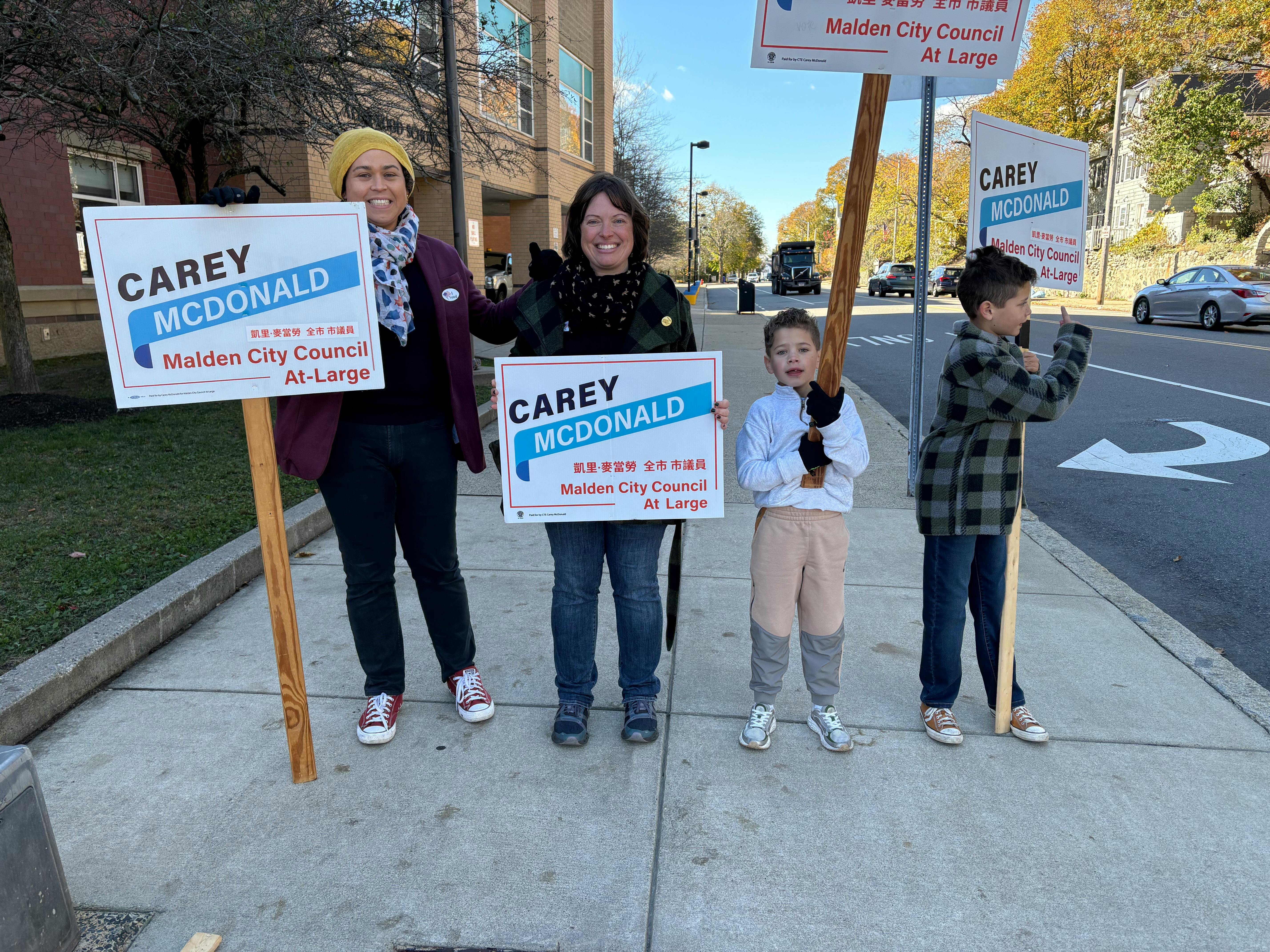 Carey, Sarah and their kids stand on the sidewalk holding Carey for City Council campaign signs, and waving to cars