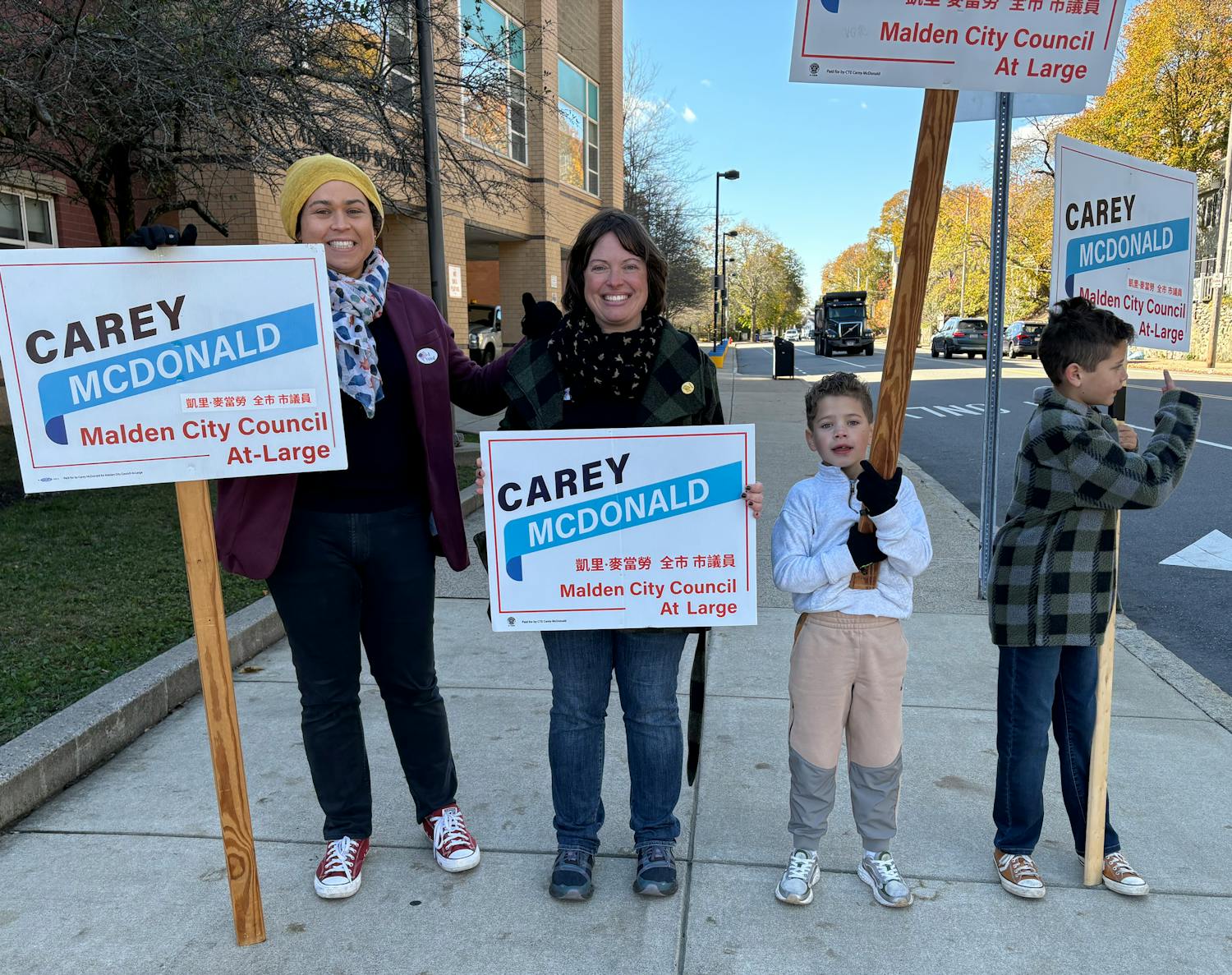 Carey, Sarah and their kids stand on the sidewalk holding Carey for City Council campaign signs, and waving to cars