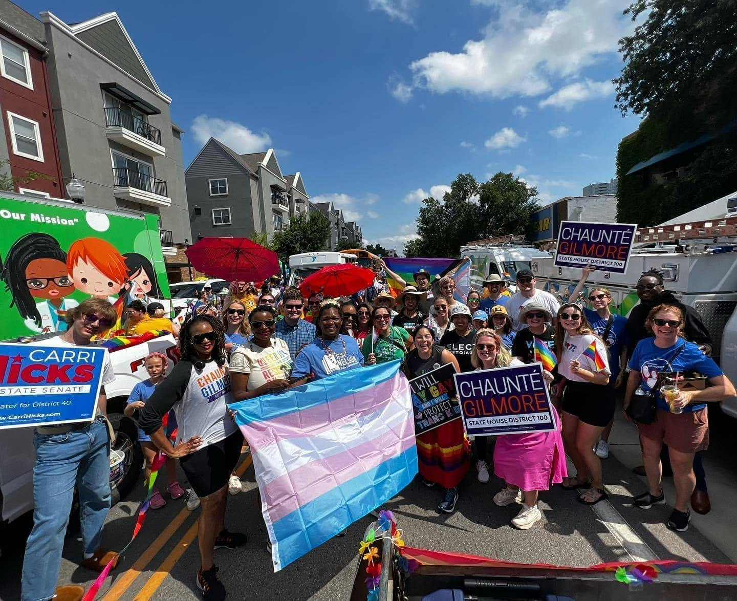 Group of Chaunté Gilmore supporters standing with her at Pride holding her signs as well as rainbow and trans flags and red umbrellas.