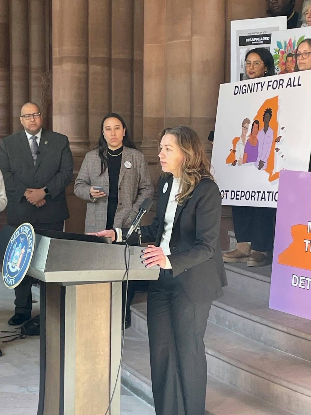 Sarah at NYS Senate podium during a press conference with activists behind, holding signs in support of Dignity Not Detention legislation
